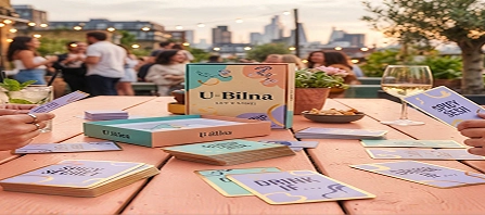 Jogo de cartas U Bilna disposto numa mesa de madeira durante uma festa no terraço ao pôr do sol, com pessoas a conversar ao fundo